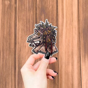 May include: A die-cut sticker featuring a stylized illustration of a horse skull adorned with a festive arrangement of holly leaves, pinecones, and multiple small golden bells tied with a dark red ribbon, finished with a decorative lace trim.