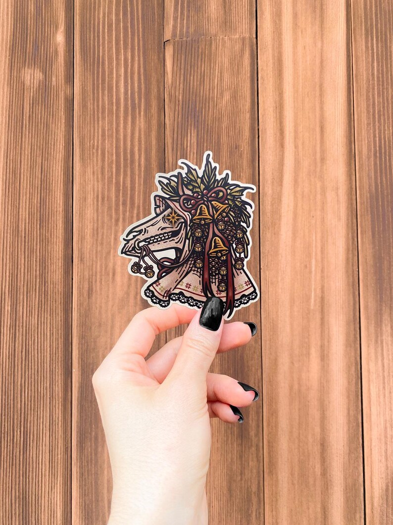 May include: A die-cut sticker featuring a stylized illustration of a horse skull adorned with a festive arrangement of holly leaves, pinecones, and multiple small golden bells tied with a dark red ribbon, finished with a decorative lace trim.