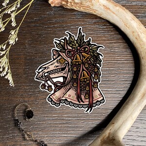 May include: A sticker featuring a horse skull adorned with a festive wreath of holly, pinecones, and golden bells. The skull has a star-shaped eye, a bridle with small bells, and a decorative fabric covering with a lace trim and snowflake pattern.