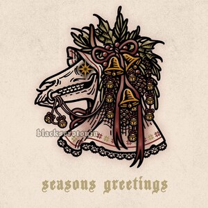 May include: A graphic illustration of a skeletal animal head wearing a bonnet adorned with holly, pinecones, and gold bells. The text blackserotonin appears near the neck, and the text seasons greetings is printed in a gothic font below the image.