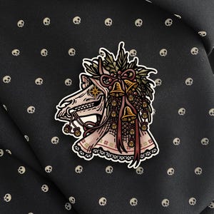 May include: A sticker featuring a stylized illustration of a horse skull adorned with a festive wreath of holly, pinecones, and golden bells. The skull has a star-shaped eye and is draped in a patterned fabric with a lace trim at the bottom edge.
