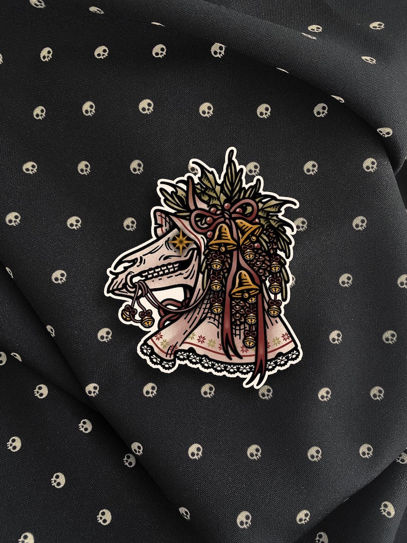 May include: A sticker featuring a stylized illustration of a horse skull adorned with a festive wreath of holly, pinecones, and golden bells. The skull has a star-shaped eye and is draped in a patterned fabric with a lace trim at the bottom edge.