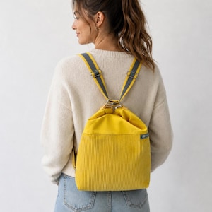 Könnte beinhalten: Ein leuchtend gelber Kordelzug-Rucksack aus Cord mit einem glatten oberen Teil aus gelbem Stoff, einem strukturierten unteren Teil aus Cord und verstellbaren grauen Trägern mit silberfarbenen Metallbeschlägen. Ein kleines blaues Markenetikett ist seitlich sichtbar.
