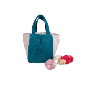 Könnte beinhalten: Eine Tragetasche aus blaugrünem Kord mit zwei passenden Henkeln und Seitenteilen mit hellrosa Blumenmuster. Ein kleiner blauer Druckknopf ist oben in der Mitte sichtbar. Neben der Tasche liegt ein mehrfarbiges Garnknäuel in Rosa, Weiß und Rot.