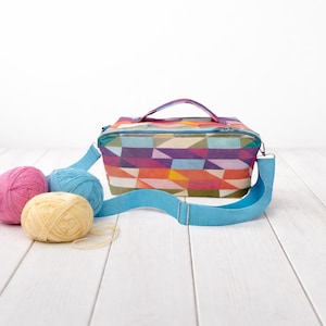 Könnte beinhalten: Eine farbenfrohe Handwerkstasche mit geometrischem Muster, einem Griff und einem blauen Schultergurt. Drei Knäuel Wolle in Rosa, Blau und Gelb liegen neben der Tasche. Die Tasche ist für Strick- oder Häkelzubehör gedacht.