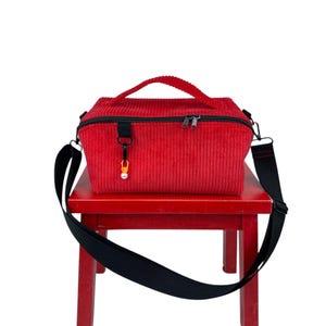 Könnte beinhalten: Eine rote Cordtasche mit schwarzem Schultergurt und oberem Griff. Die Tasche hat einen Reißverschluss und einen kleinen Anhänger an der Seite. Sie steht auf einem roten Hocker.