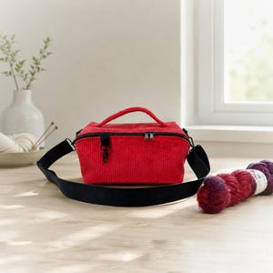 Könnte beinhalten: Eine rechteckige Tasche aus rotem Kord mit einem Tragegriff, einem schwarzen Reißverschluss und einem breiten, verstellbaren schwarzen Schultergurt. Die Tasche verfügt über einen kleinen schwarzen Metallclip an der Vorderseite.