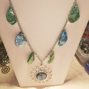 May include: A silver chain necklace with a sun pendant featuring a blue and green iridescent shell. The necklace has several smaller shell charms in shades of blue and green.