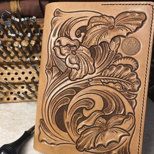 Può includere: Un diario in pelle marrone con un'intricata lavorazione floreale. La copertina presenta un design dettagliato di fiori e foglie, con un motivo a spirale al centro. Il diario è chiuso e ha un bordo cucito.