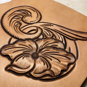 May include: A close-up of a brown leather piece with a carved design of a flower and a wave. The carving is detailed and intricate.