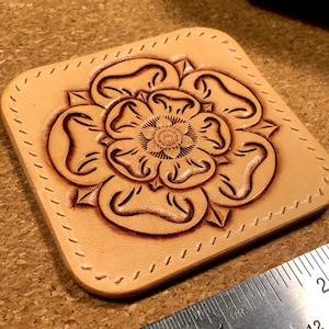 May include: A square leather coaster featuring a detailed, carved rose design in brown. The coaster has a stitched border.