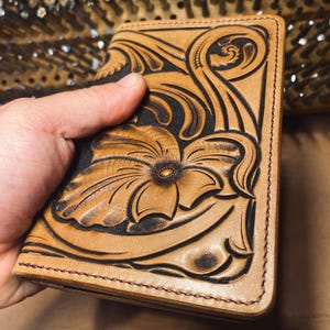 May include: A tan leather wallet with a floral design. The wallet features a detailed embossed flower and leaf pattern with dark brown accents. The edges are stitched with brown thread, and the wallet is held in a hand.