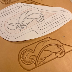 Könnte beinhalten: Ein Lederhandwerksprojekt mit einem Federdesign. Eine Papierform mit dem Design liegt auf dem Leder. Das Leder wurde mit dem Federdesign geprägt und die Initialen "J.P.C." befinden sich in einem Kreis unten links.