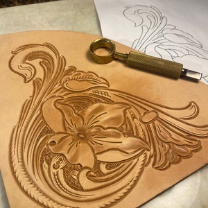 May include: A close-up of a leather craft project in progress. A floral design is carved into the light brown leather. A brass swivel knife and a design template are visible.