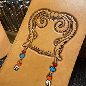 May include: A brown leather journal cover with a tooled design of a decorative frame with two dangling strands of beads in blue, orange, and red.