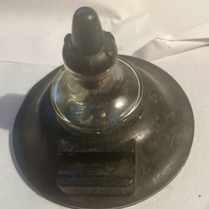 May include: A vintage desk inkwell featuring a circular black base with a built-in pen rest and a central clear glass reservoir topped with a black cap. The base has a wide, flat design with a smooth finish, providing a stable foundation for the glass ink container.