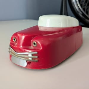 Könnte beinhalten: Ein rotes Spielzeugauto aus Kunststoff mit weißem Dach, das zwei kleine kreisförmige Metallakzente als Augen auf der Motorhaube, einen silberfarbenen Metallkühlergrill mit drei horizontalen Streben und ein glattes, abgerundetes Design aufweist.