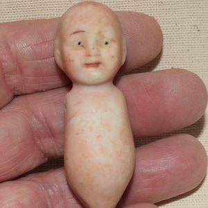 May include: A small, antique porcelain doll with a bald head, painted facial features, and a pale pink body. The doll is missing an arm. The doll is held in a hand.
