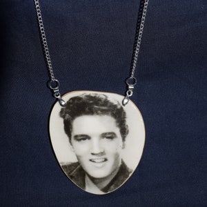 May include: A pendant necklace with a black and white image of Elvis Presley. The pendant is a light beige colour and is suspended from a silver chain. The background is a dark blue.