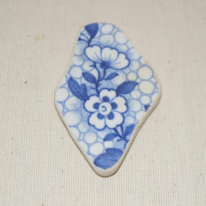 May include: A blue and white ceramic shard with a floral design. The shard is diamond-shaped, featuring a blue floral pattern set against a backdrop of white circles. The design includes flowers, leaves, and stems.