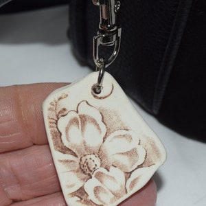 May include: A cream-coloured, irregular-shaped keychain pendant with a floral design in brown. The pendant is attached to a silver-coloured metal clasp. The floral design features a large flower with detailed petals.
