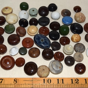 May include: A selection of vintage cabinet knobs in diverse colours and materials, including ceramic, wood, and stone. The knobs are round and in shades of brown, white, green, and blue. A wooden ruler is at the bottom.