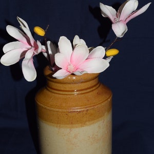 May include: A cylindrical ceramic vase in shades of brown and cream, containing magnolia flowers. The flowers have white petals with pink centres and yellow buds. The vase has a textured surface and is approximately 20 cm tall.