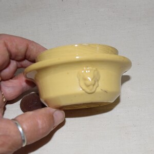 May include: A small, yellow ceramic bowl with a lion's head relief. The bowl has a rounded shape with a flat rim and a flared base. The bowl is held in a hand. The bowl is likely vintage.