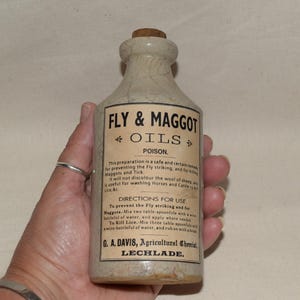 May include: Vintage ceramic bottle marked "FLY & MAGGOT OILS POISON." The bottle features a cork stopper and a cream-colored finish. The label provides instructions for use and the name G. A. Davis, Agricultural Chemist, Lechlade. The bottle is for livestock.