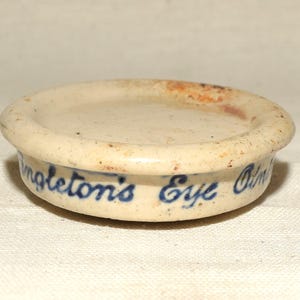 May include: A small, cream-coloured ceramic lid with a raised edge. The lid has a flat top and a slightly curved side. The words "Singleton's Eye Ointment" are written in blue script around the side.