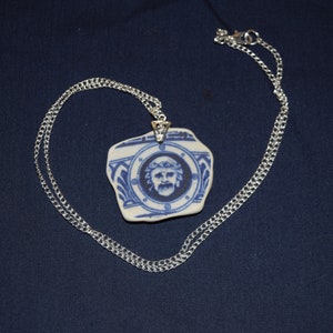 May include: A silver chain necklace with a blue and white ceramic pendant. The pendant depicts a man's portrait. The necklace is presented against a dark blue backdrop.