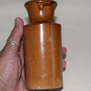 May include: A brown ceramic jar with a cylindrical body and a flared neck. The jar has a glossy finish and appears to be handmade. The jar is held in a person's hand.