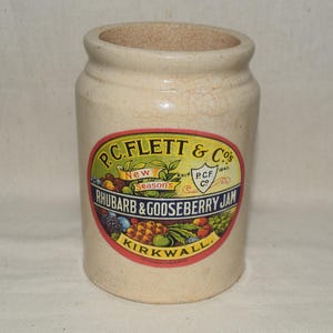 May include: An antique stoneware jar in a cream colour with a cracked surface. The jar has a colourful label reading "P.C. Flett & Co's Rhubarb & Gooseberry Jam, Kirkwall." The label features fruit illustrations.