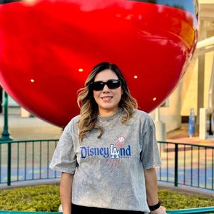 May include: A person wearing sunglasses and a gray t-shirt with the text "Disneyland Resort" in blue and red. A large red and black Mickey Mouse head is in the background.