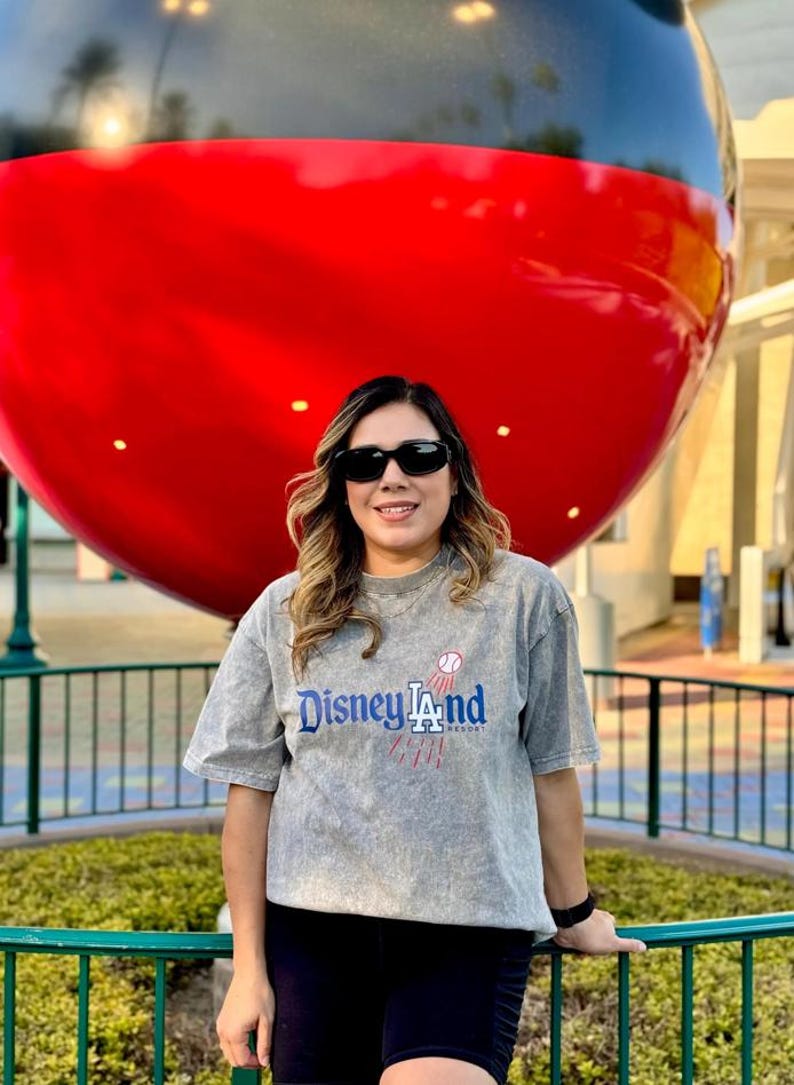 May include: A person wearing sunglasses and a gray t-shirt with the text "Disneyland Resort" in blue and red. A large red and black Mickey Mouse head is in the background.