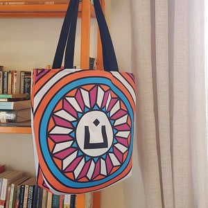 Puede incluir: Una bolsa de tela blanca con asas negras. La bolsa tiene un diseño circular colorido con un patrón geométrico en tonos naranja, rosa, azul y blanco. El centro del círculo presenta un símbolo negro.
