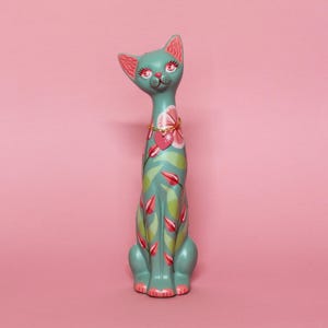 May include: A teal cat figurine with pink accents and floral designs. The cat has a gold necklace with a heart pendant and is decorated with pink and green leaves and flowers. The cat is sitting upright on a pink background.
