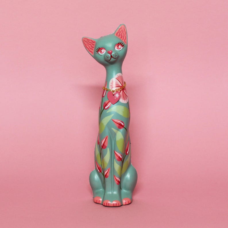 May include: A teal cat figurine with pink accents and floral designs. The cat has a gold necklace with a heart pendant and is decorated with pink and green leaves and flowers. The cat is sitting upright on a pink background.