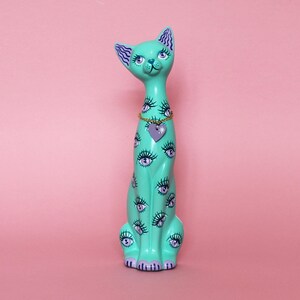 May include: A tall, turquoise cat figurine with a whimsical design. The cat is adorned with multiple eye motifs and a purple heart pendant. The cat has a gold chain necklace and is set against a pink background.