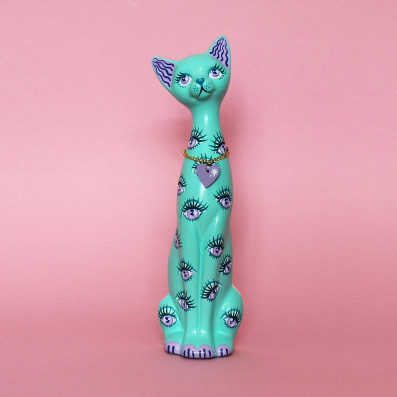 May include: A tall, turquoise cat figurine with a whimsical design. The cat is adorned with multiple eye motifs and a purple heart pendant. The cat has a gold chain necklace and is set against a pink background.