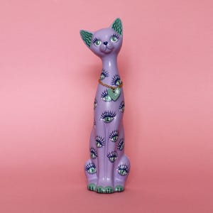 May include: A lavender cat figurine with multiple eye designs. The cat has green accents on its ears, paws, and eyes. A gold chain necklace with a heart-shaped pendant hangs around its neck. The figurine is set against a pink background.