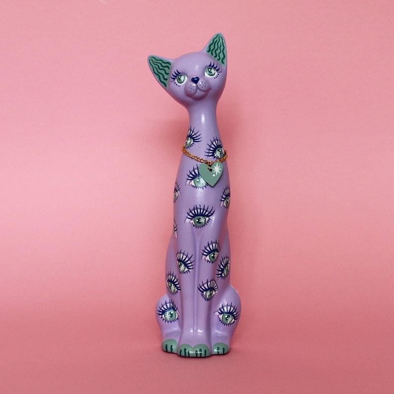 May include: A lavender cat figurine with multiple eye designs. The cat has green accents on its ears, paws, and eyes. A gold chain necklace with a heart-shaped pendant hangs around its neck. The figurine is set against a pink background.