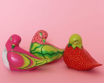 Puede incluir: Tres figuras de pájaros coloridos. Una es rosa con detalles amarillos y lunares blancos, otra tiene un diseño de sandía y la tercera es una fresa roja. Todas sobre un fondo rosa.