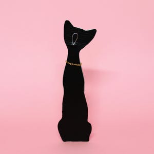 May include: A black cat-shaped jewelry display stand with a gold-colored chain necklace. The stand is made of a dark material and has a simple design. The background is a solid pink color.