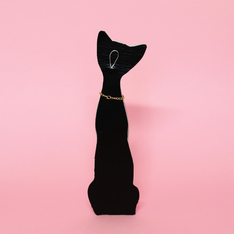May include: A black cat-shaped jewelry display stand with a gold-colored chain necklace. The stand is made of a dark material and has a simple design. The background is a solid pink color.