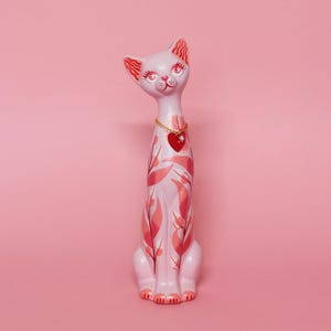 May include: A light pink ceramic cat figurine with a gold chain necklace and a red heart pendant. The cat is decorated with pink leaf patterns and has red accents on its ears and face. The figurine is approximately 12 inches tall.