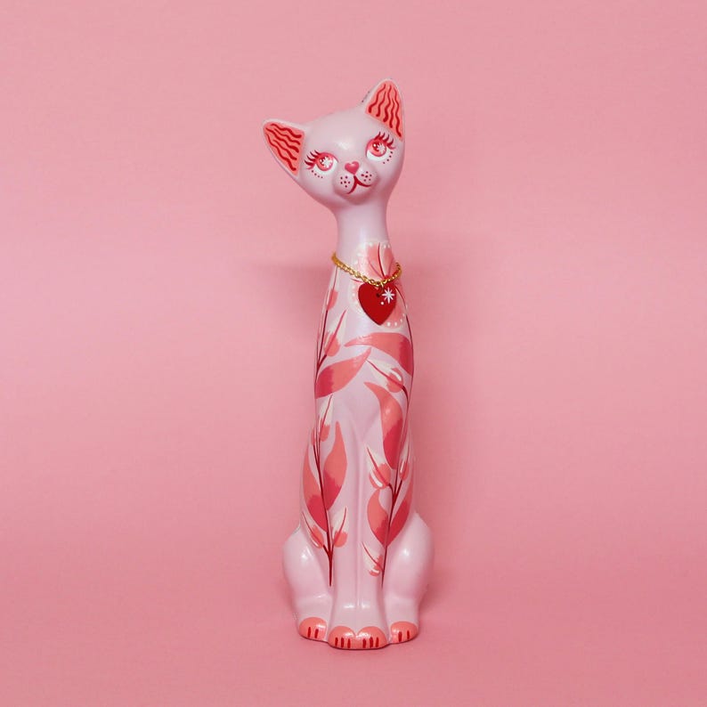May include: A light pink ceramic cat figurine with a gold chain necklace and a red heart pendant. The cat is decorated with pink leaf patterns and has red accents on its ears and face. The figurine is approximately 12 inches tall.