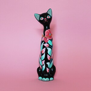 May include: A black cat figurine with a floral design in pink, teal, and white. The cat has a pink flower collar and teal accents on its ears. The figurine is set against a pink background.
