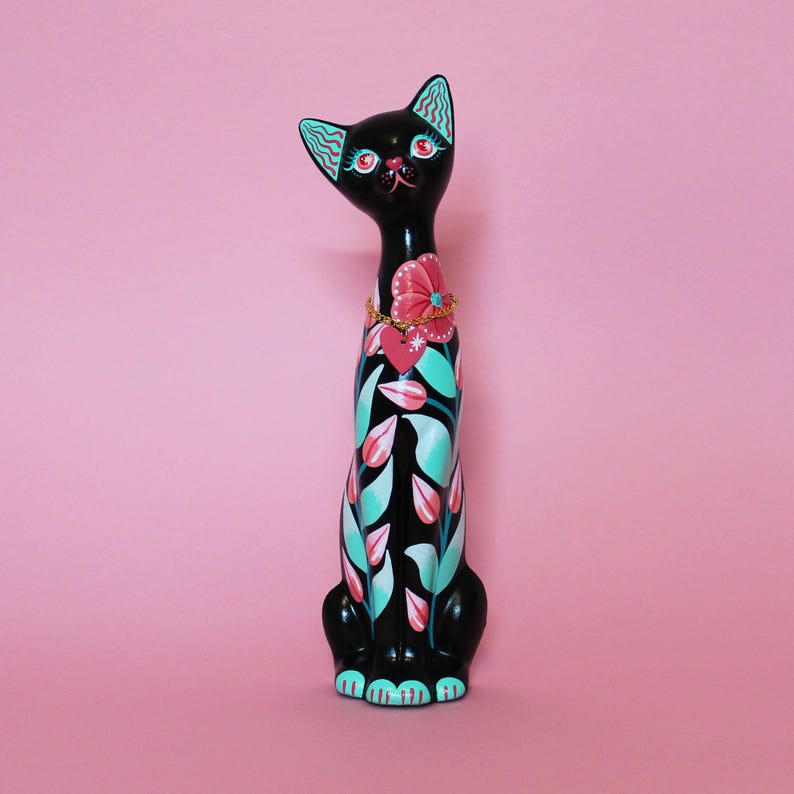 May include: A black cat figurine with a floral design in pink, teal, and white. The cat has a pink flower collar and teal accents on its ears. The figurine is set against a pink background.