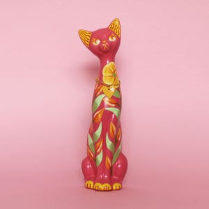 May include: A tall, pink cat figurine with yellow and green floral designs. The cat has yellow ears and a yellow flower necklace with a heart pendant. The cat's paws are yellow, and the background is pink.